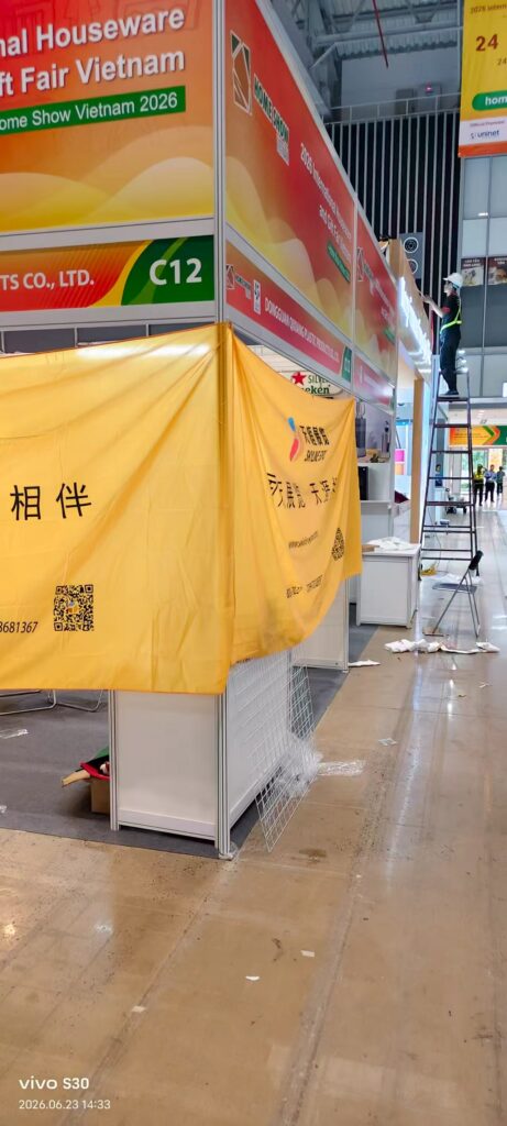Home Show Vietnam 2026布展现场照片,展馆内黄色展架整齐排列,展架上摆放着各类家居礼品样品,展位上方悬挂银色装饰品牌标识,背景可见其他展商正在布展。
